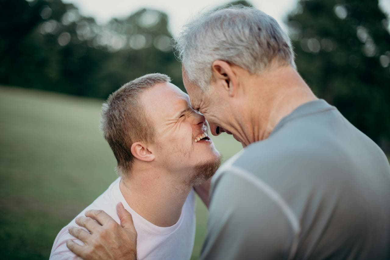 Two men laughing together outdoors, foreheads touching.