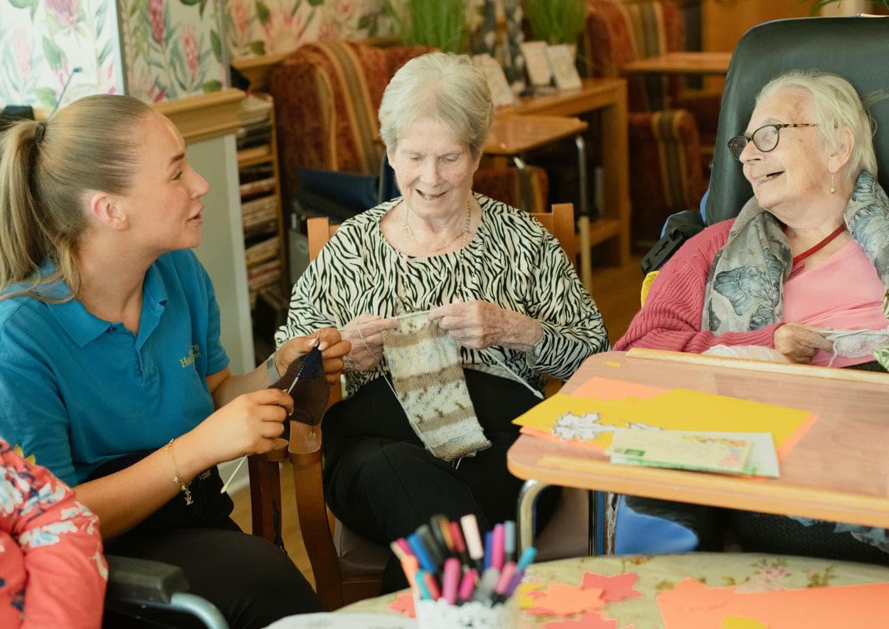 A support worker and two women knitting together in a care home lounge.